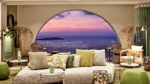 A cozy lounge area with a large arched window offering a scenic view of the sea and a sunset. - Myconian Korali Relais & Chateaux (Mykonos)