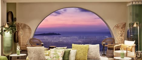 A cozy lounge area with a large arched window offering a scenic view of the sea and a sunset.