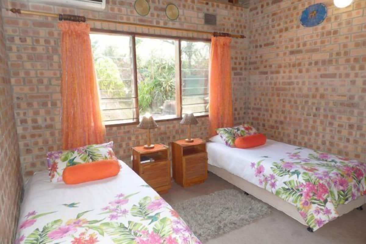 A room with two beds, a window, a wall clock, and a small table with a lamp.