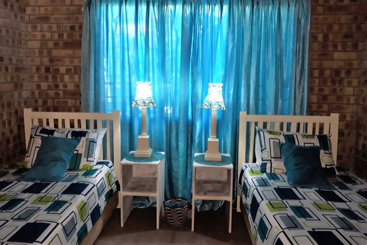 A room with two beds, each with patterned bedding, a brick wall, and a blue curtain.