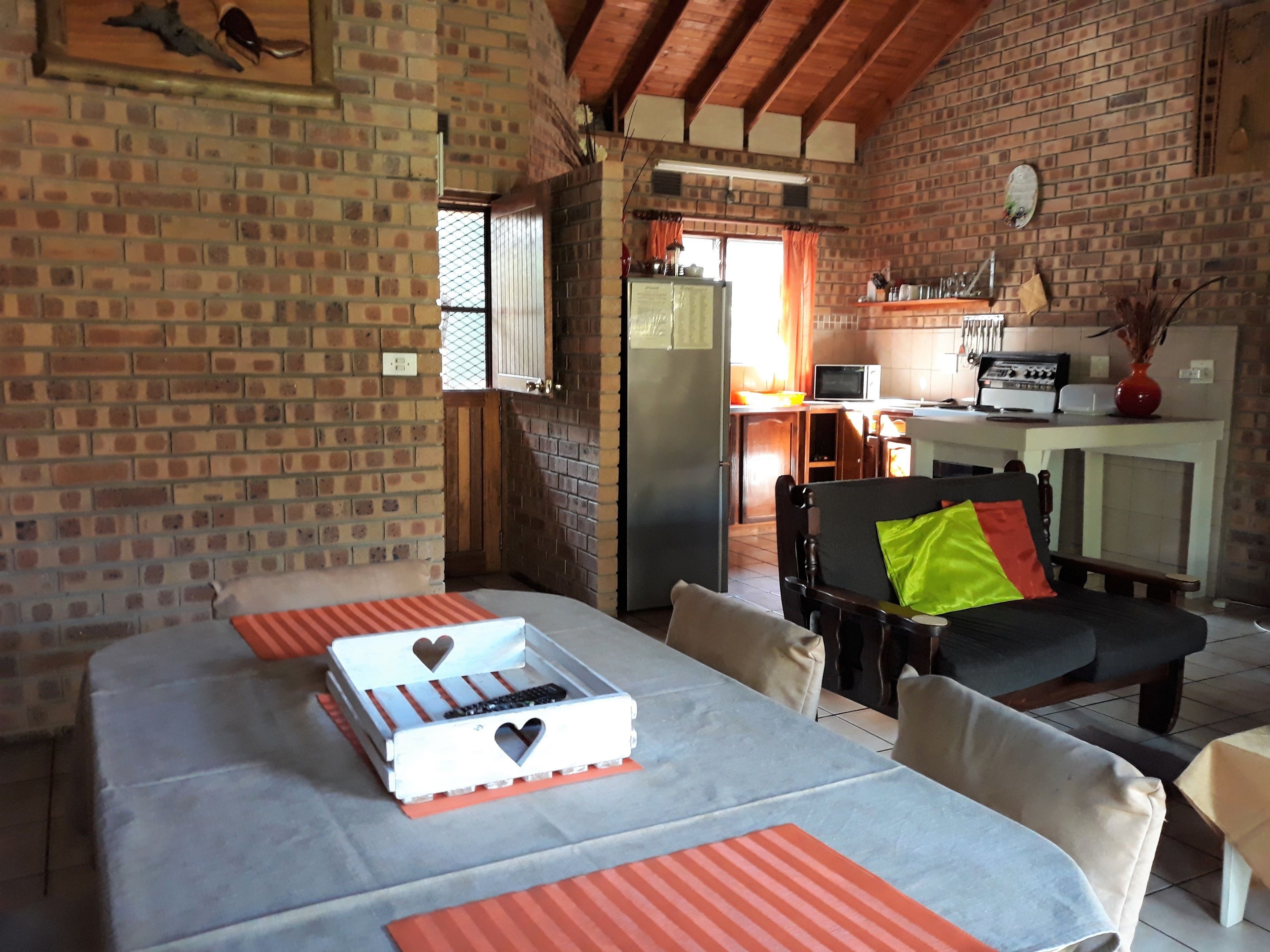 Una habitación acogedora con pared de ladrillo, una mesa de comedor con manteles individuales anaranjados, una silla negra con un cojín verde, y una zona de cocina al fondo.