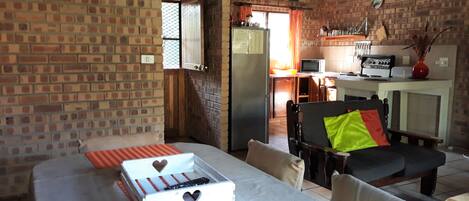 Una habitación acogedora con pared de ladrillo, una mesa de comedor con manteles individuales anaranjados, una silla negra con un cojín verde, y una zona de cocina al fondo.
