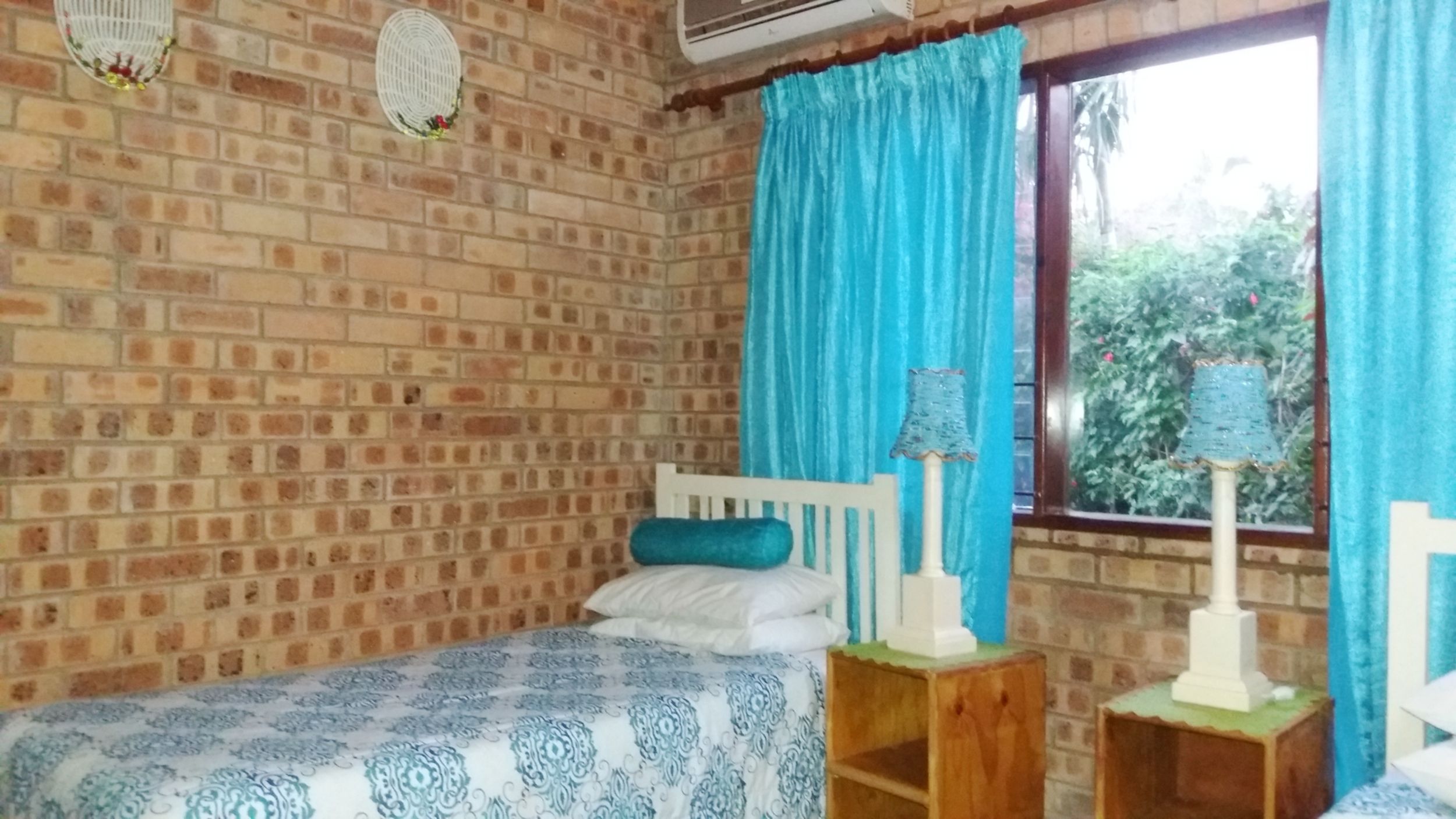 Una habitación con pared de ladrillo, cama con colcha estampada, cabecera blanca, cortina azul y ventana con vistas a la vegetación.