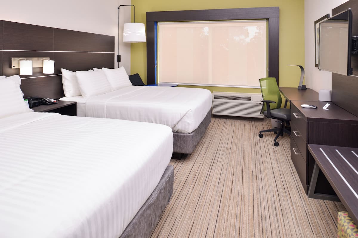 A hotel room with two beds, a desk, a chair, a lamp, and a large window with blinds.