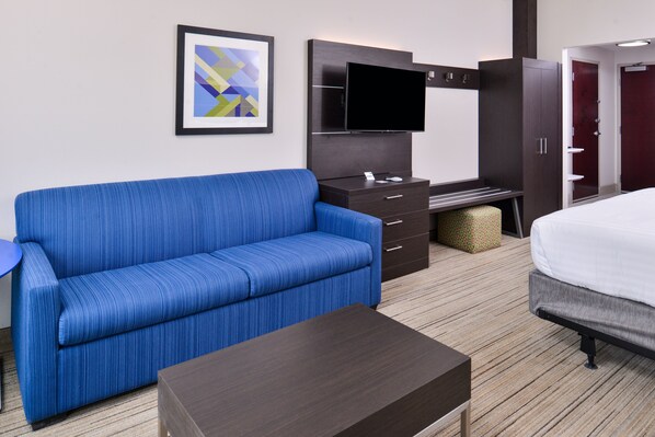 Quarto de hotel com sofá azul, televisão de tela plana, cama, mesa de centro de madeira escura e uma obra de arte abstrata emoldurada na parede.