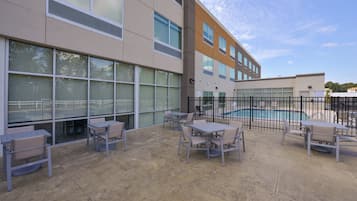 An outdoor patio area with tables and chairs, a swimming pool, and a building with large windows.