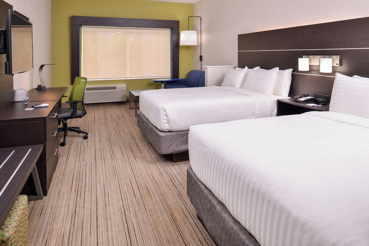 A hotel room with two beds, a desk with a chair, a small table, and a window with blinds.