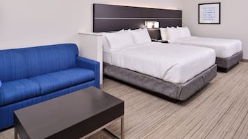 A hotel room with a blue sofa, two beds with white linens, a dark wooden coffee table, and a headboard with wall-mounted lights.
