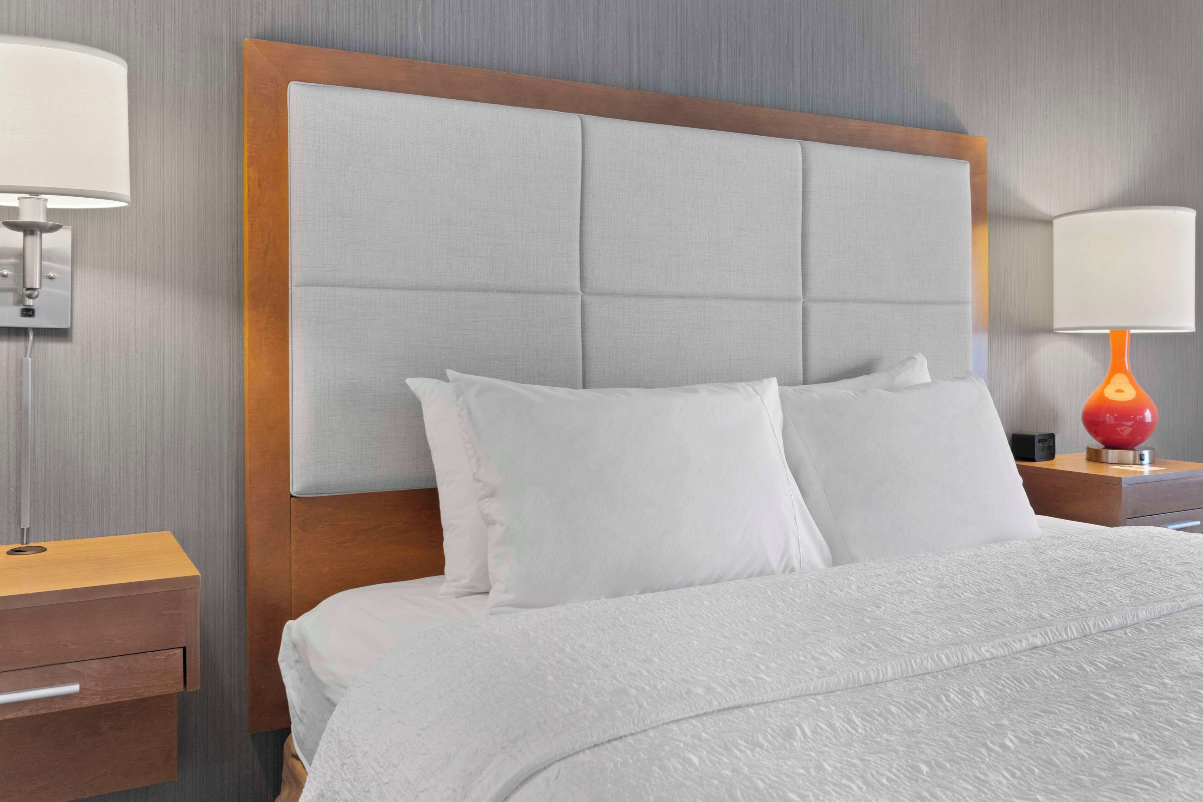 Uma cama bem arrumada com cabeceira acolchoada, um criado-mudo de madeira com uma luminária e um abajur vermelho na mesinha.
