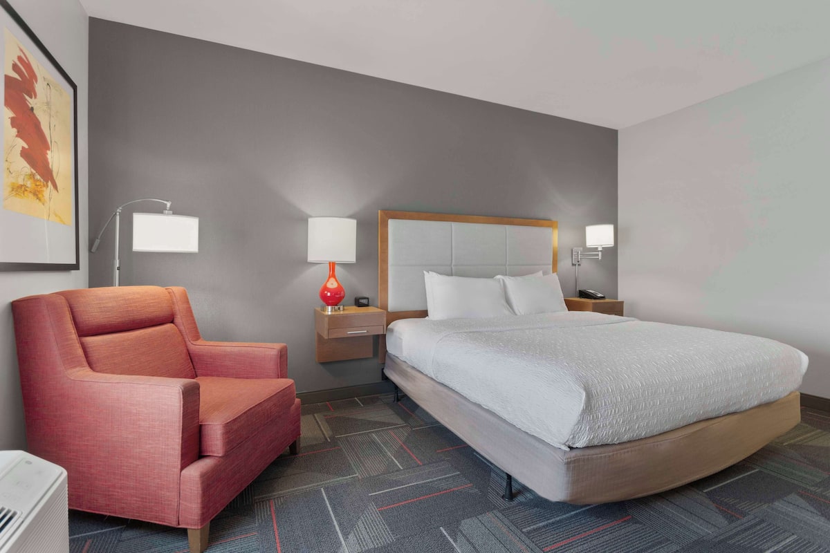 A hotel room with a large bed, a red armchair, two bedside tables with lamps, and a framed artwork on the wall.