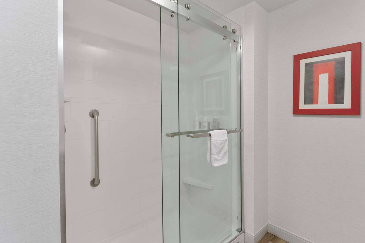 A modern bathroom with a glass shower enclosure, a white towel hanging on the door, and a framed artwork on the wall.