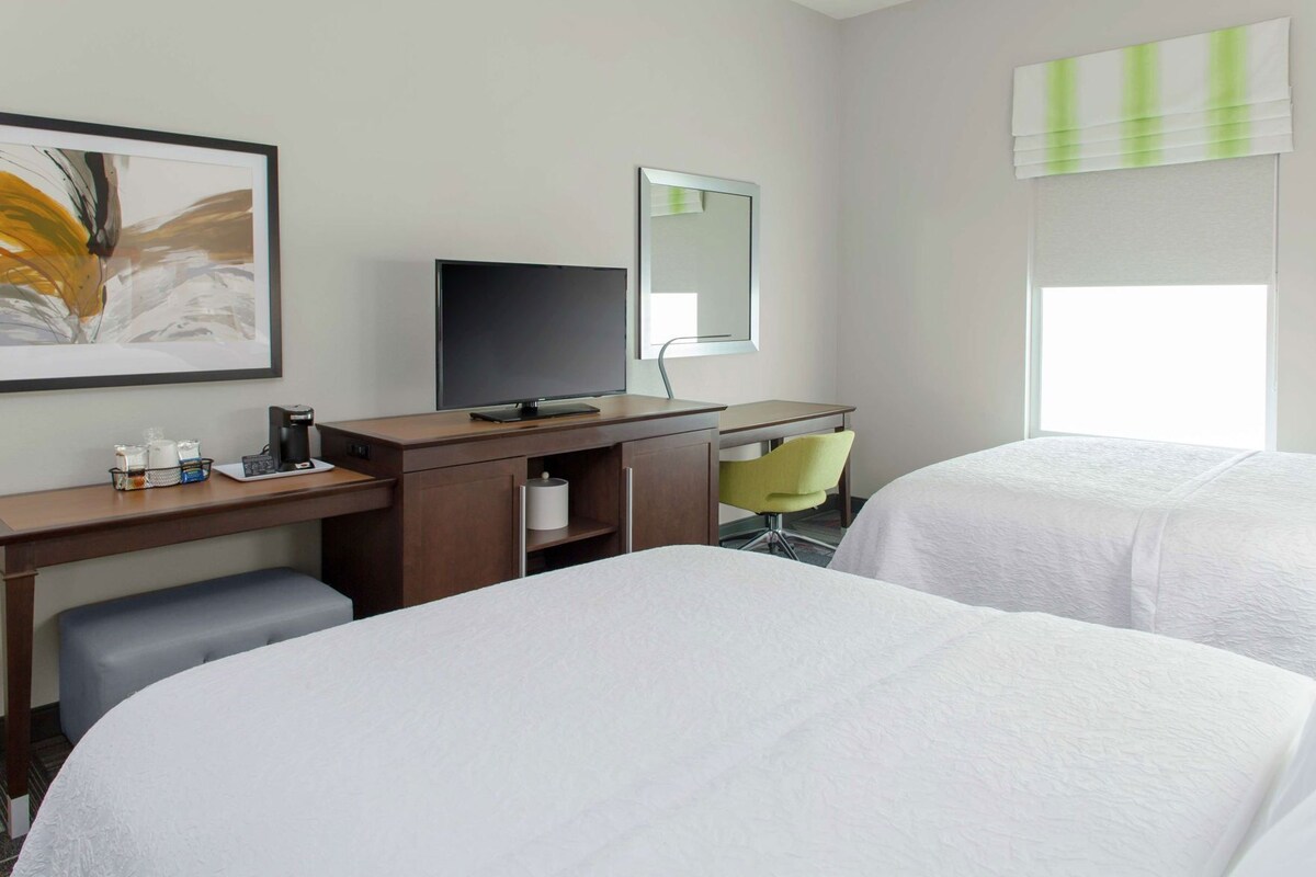 A hotel room with two beds, a desk with a TV, a chair, a mirror, and a window with blinds.