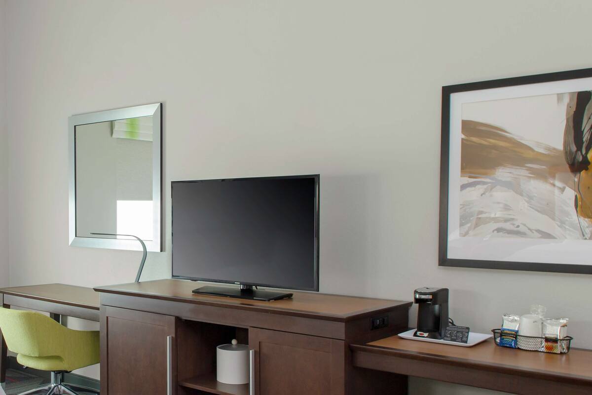 A hotel room with a desk, a flat-screen TV, a coffee maker, and a framed abstract painting on the wall.