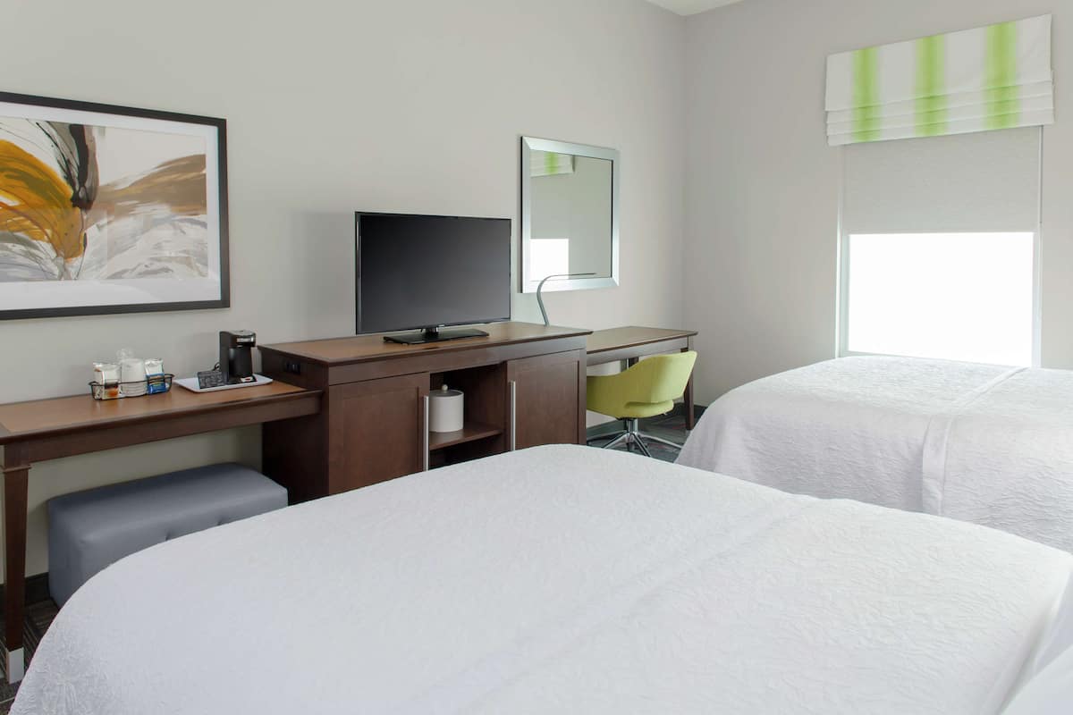 A hotel room with two beds, a desk with a computer, a chair, a mirror, and a window with blinds.