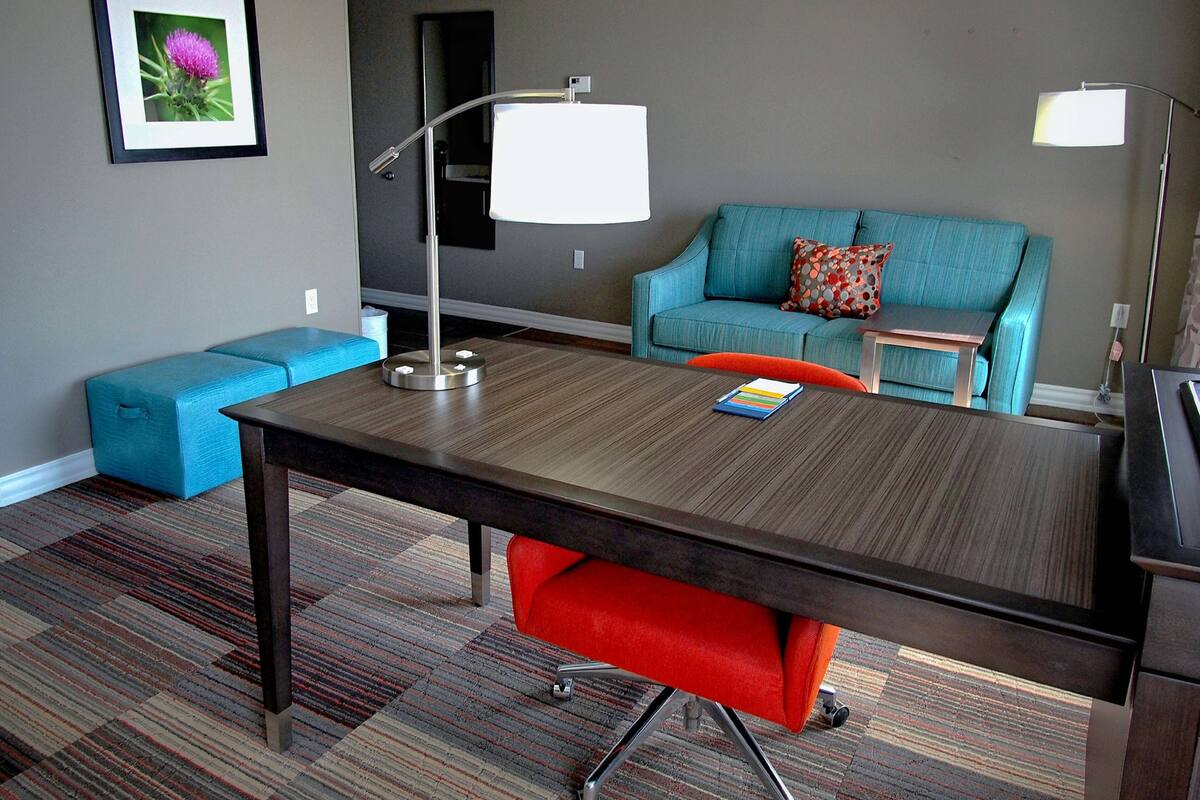 A modern hotel room with a wooden desk, a blue sofa, a small side table, and a red chair.