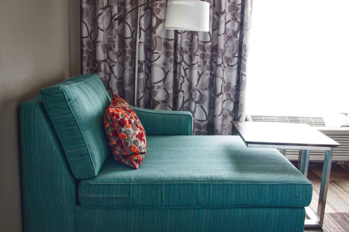 A teal sofa with a patterned pillow, a white lamp, and a patterned curtain in a room with a radiator.