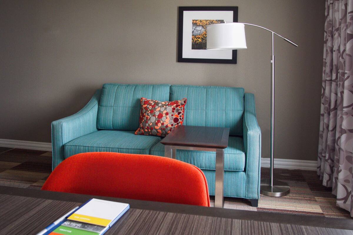 A room with a teal sofa, a small table, a red chair, a framed picture on the wall, and a floor lamp.
