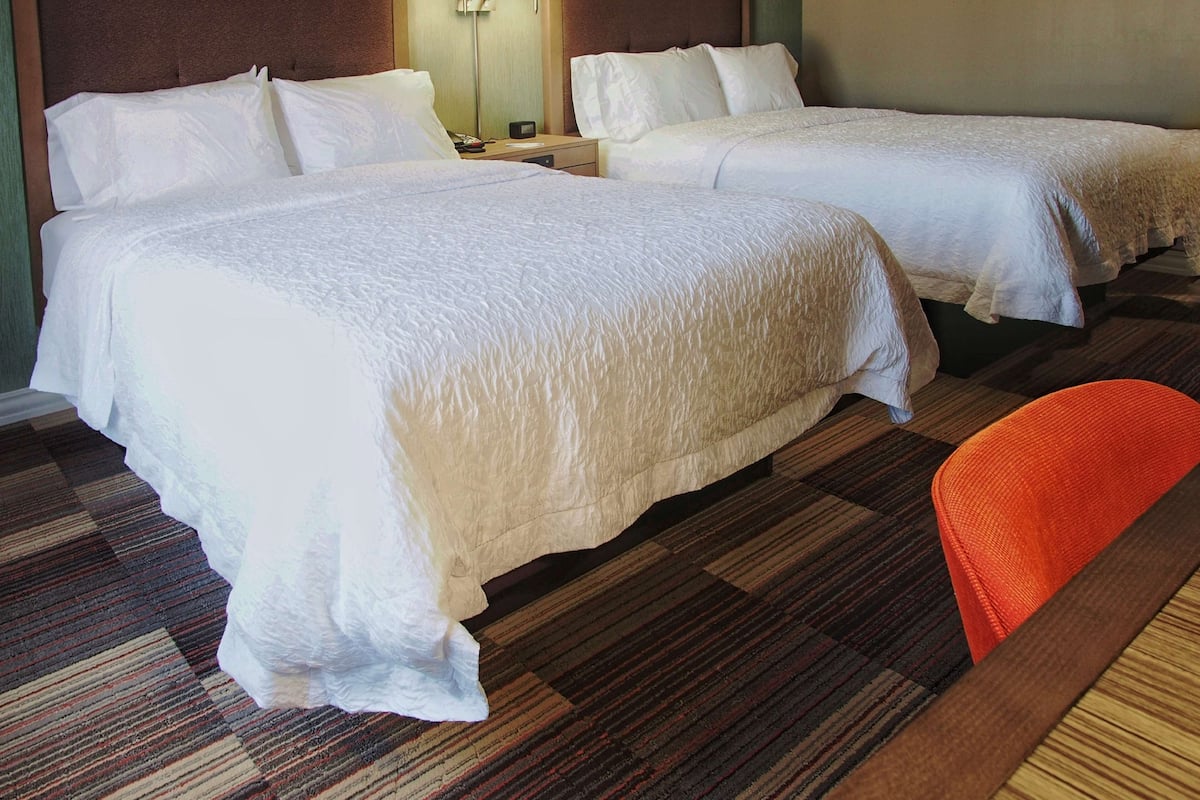 A hotel room with two beds, a nightstand, a lamp, and a chair.