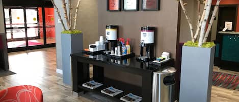 Un mostrador de recepción con máquinas de café, un cubo de basura y revistas. Hay dos plantas en macetas con ramas y una silla roja en primer plano.