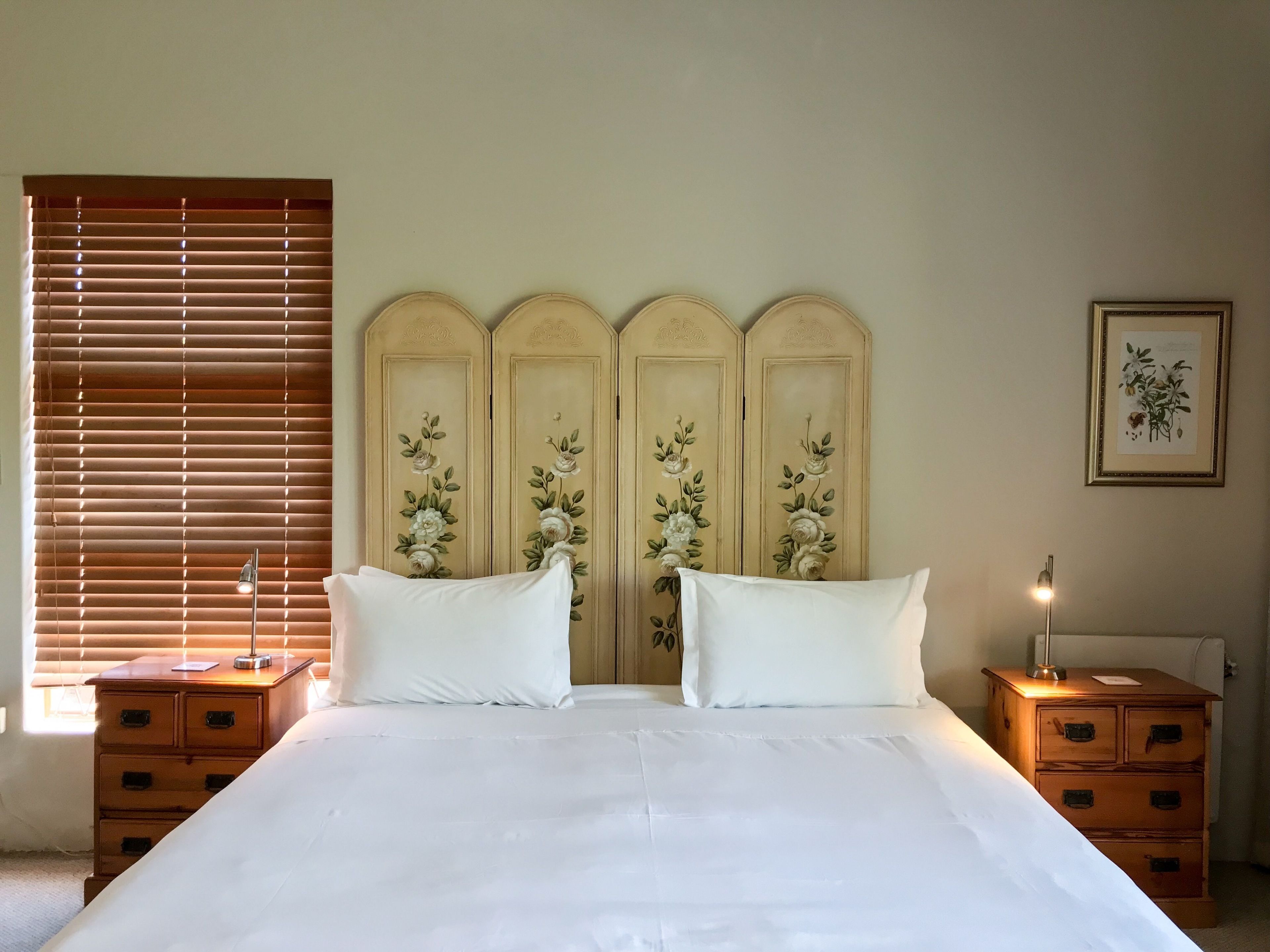 A bedroom with a large bed, two nightstands, a wooden headboard with floral designs, a window with blinds, and a framed botanical print on the wall.