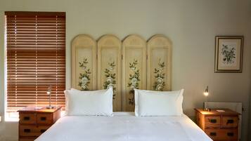A bedroom with a large bed, two nightstands, a wooden headboard with floral designs, a window with blinds, and a framed botanical print on the wall.