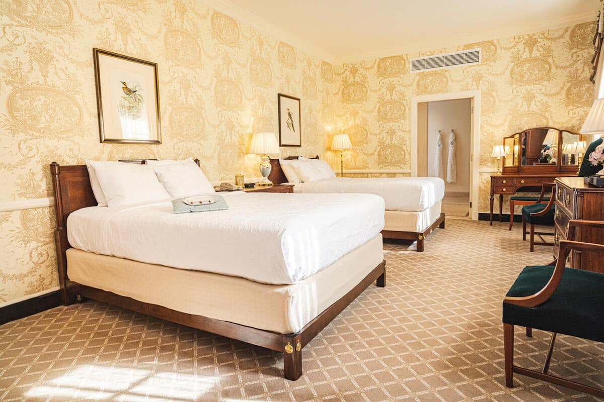 A hotel room with two beds, a desk, and a chair. There are lamps on the bedside tables and a mirror on the wall.