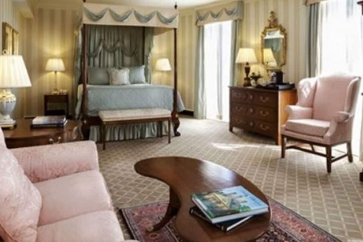A bedroom with a four-poster bed, a sitting area with a sofa and armchair, a wooden dresser, and a nightstand with a lamp.