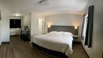A bedroom with a large bed, a desk, and a chair. There is a window with curtains, a ceiling light, and a wall-mounted lamp.
