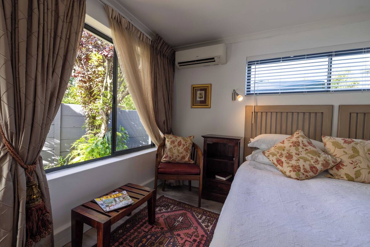 Ein Schlafzimmer mit Bett, Stuhl, einem kleinen Tisch, einem Fenster mit Vorhängen und einer Wandklimaanlage.