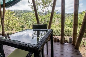 A glass-top table with a woven chair on a wooden deck, surrounded by bamboo poles, overlooking a lush green landscape.