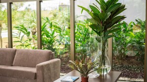 A room with a sofa, a table with a potted plant, and a view of greenery outside. - Cambuci Hotel (Camaçari)