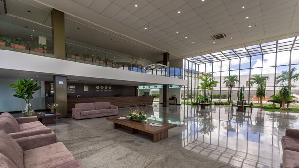 A spacious lobby with a high ceiling, a wooden reception desk, multiple seating areas, and a glass wall offering a view of the outdoors. - Cambuci Hotel (Camaçari)