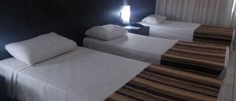Habitación de hotel con dos camas, cada una con ropa de cama y almohadas blancas. Un mesita de noche con lámpara se encuentra entre las camas.