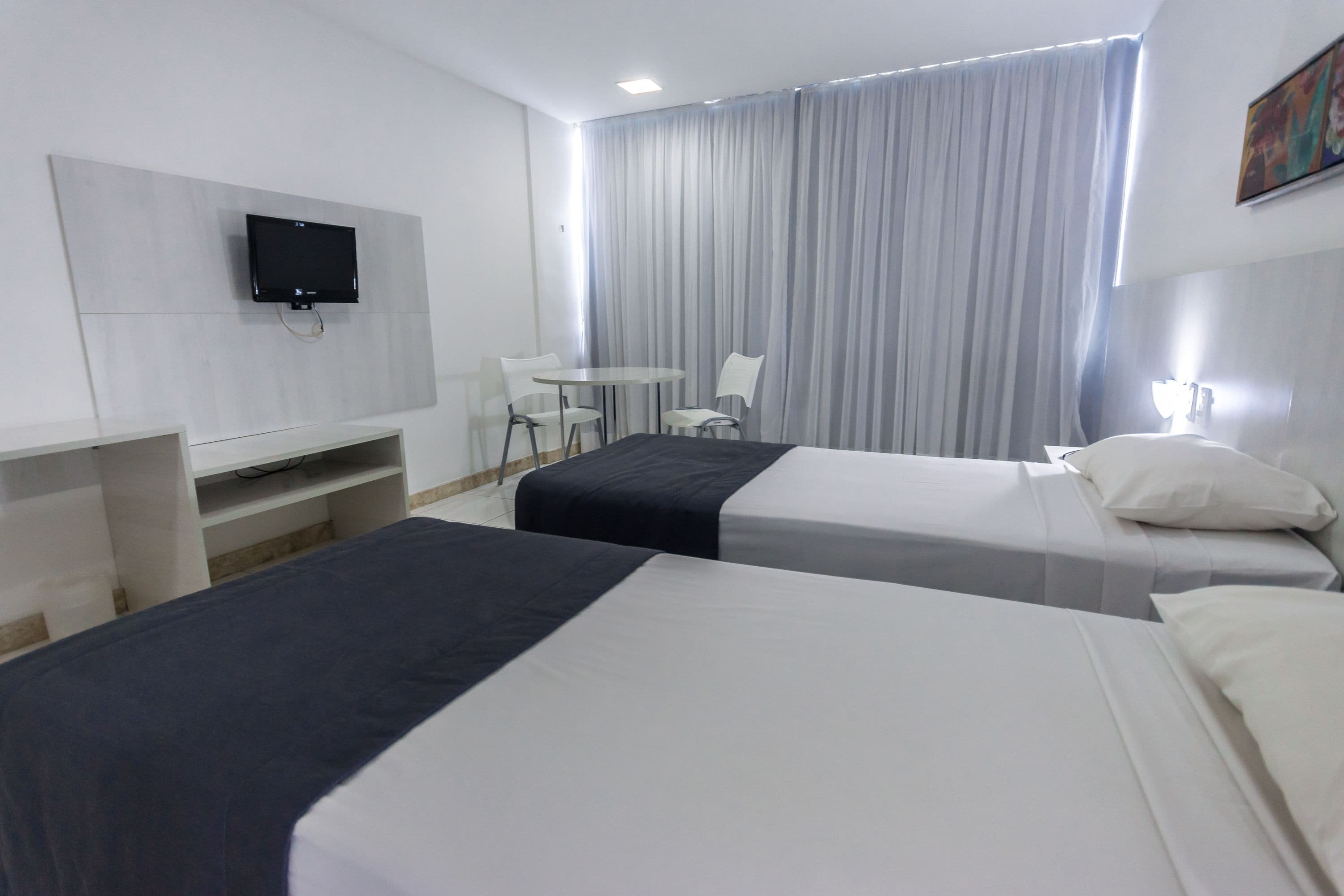 Habitación de hotel con dos camas, un televisor montado en la pared, un escritorio con silla y un ventanal con cortinas.