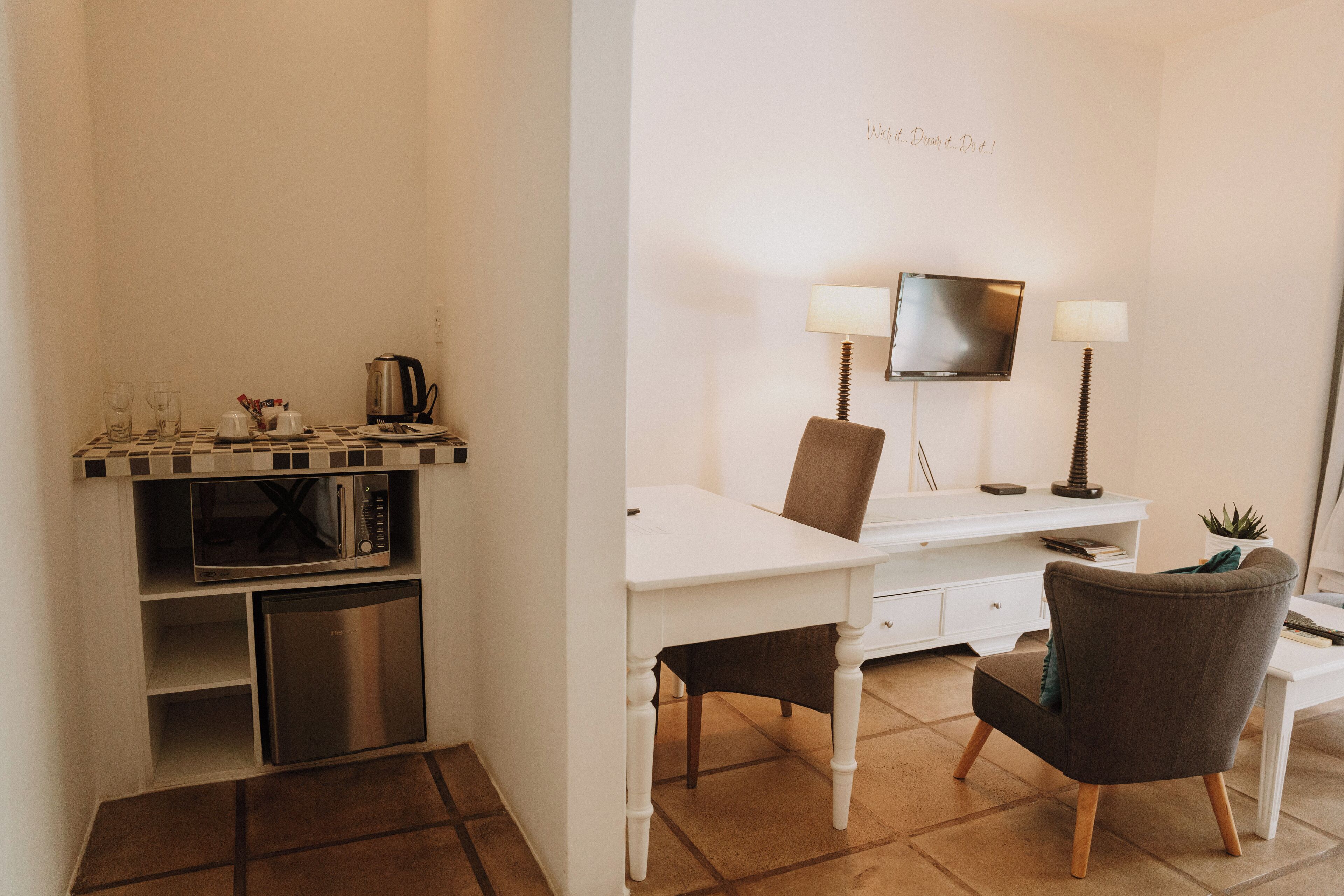 Habitación de hotel compacta con kitchenette, un escritorio con dos sillas, un televisor de pantalla plana y una pequeña planta en maceta.