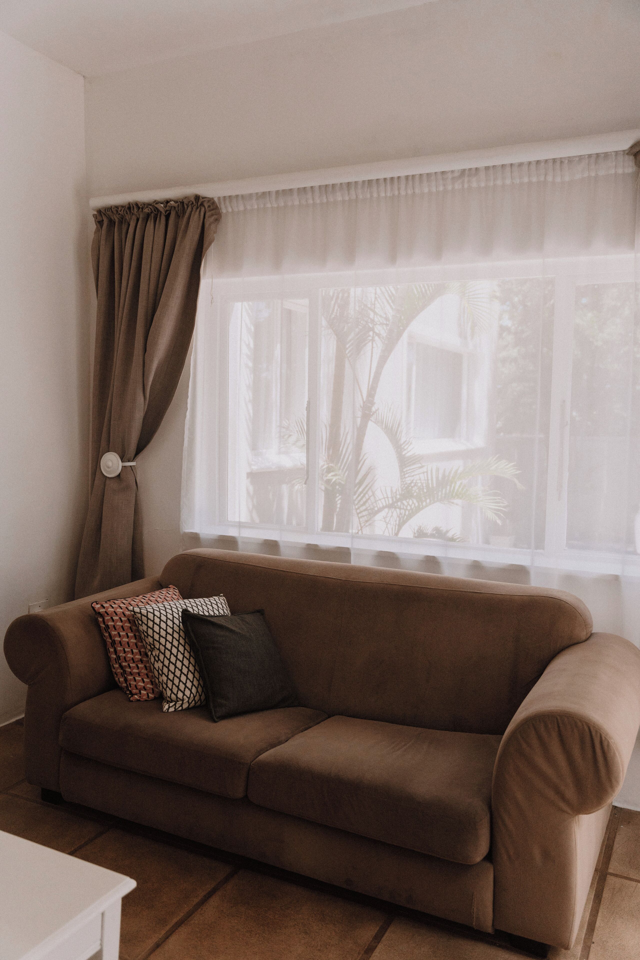 A living room with a brown sofa, a white table, and a window with sheer curtains.