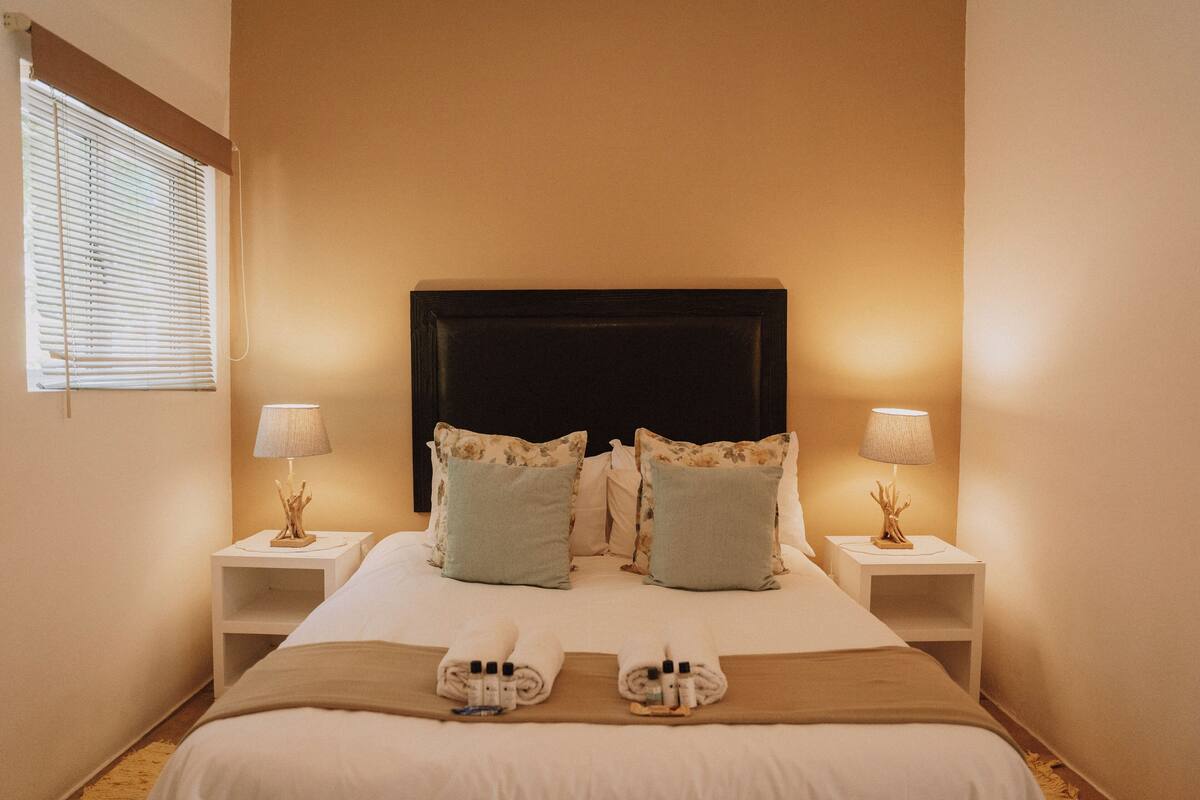 Une chambre d’hôtel avec un grand lit, deux tables de chevet avec des lampes et une fenêtre avec des stores.