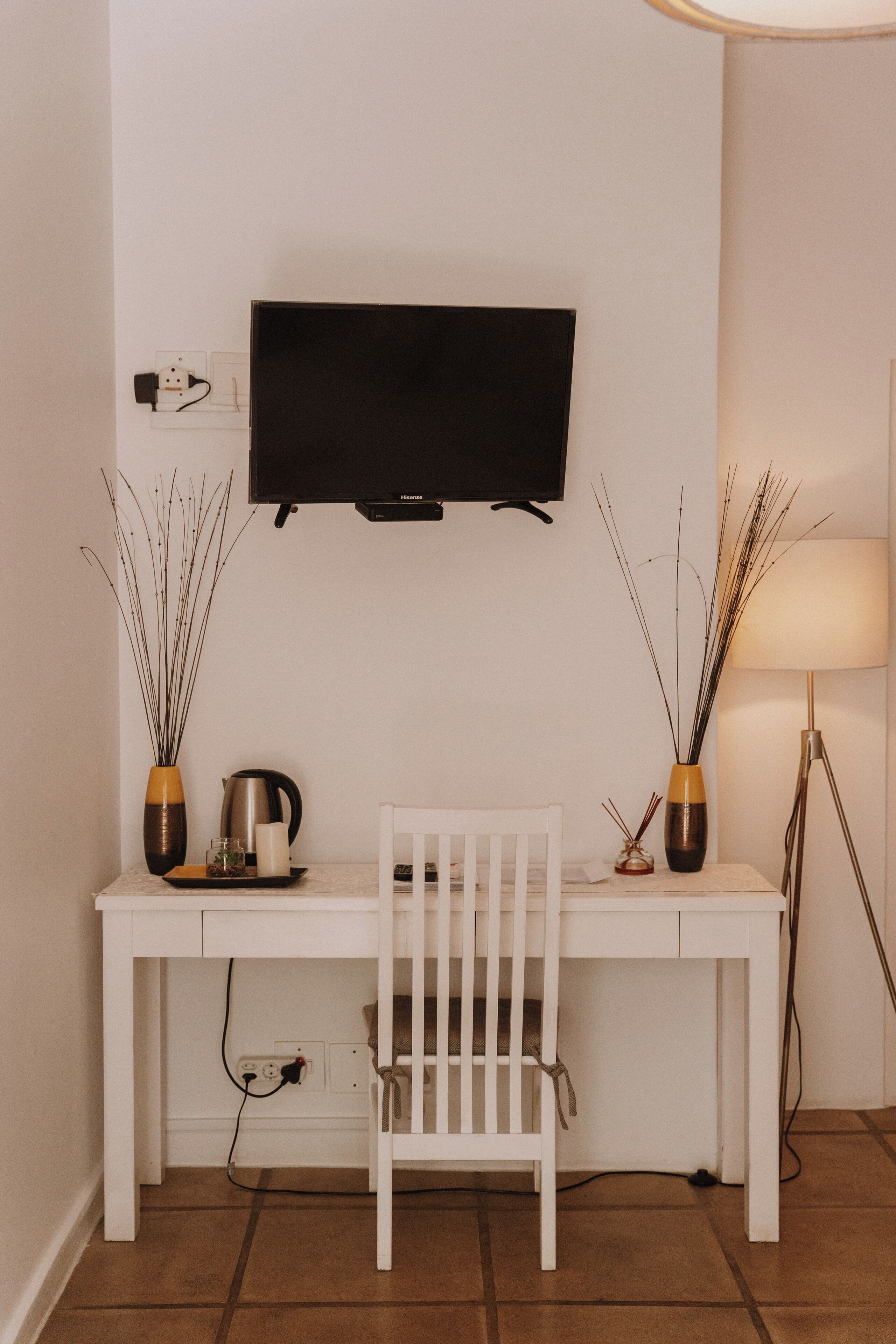 A white desk with a TV mounted above it, a white chair, two vases with decorative branches, a floor lamp, and a kettle on the desk.