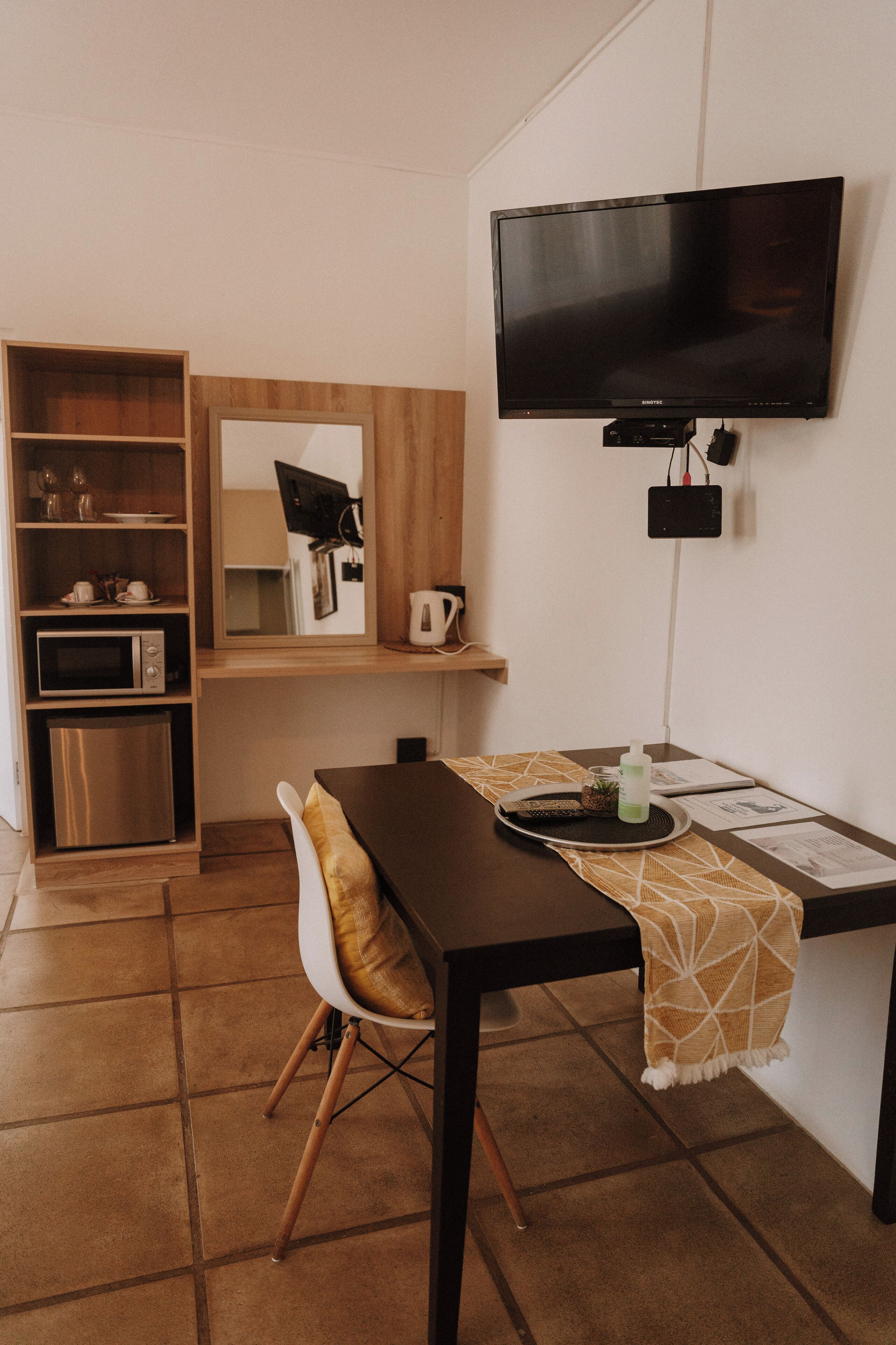 A compact kitchenette with a microwave, coffee maker, and a small refrigerator. A dining table with a chair is positioned in front of a flat-screen TV mounted on the wall.