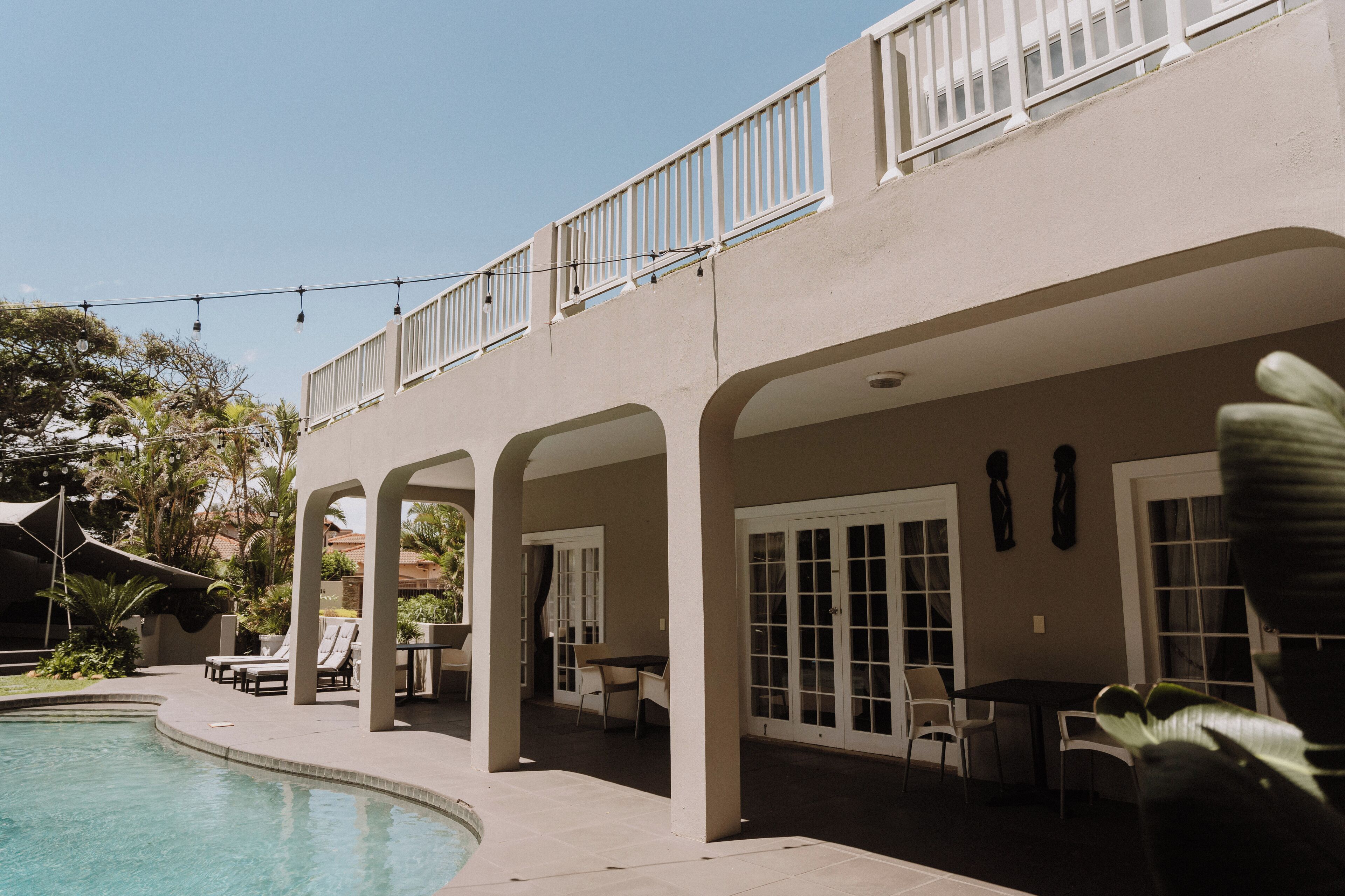 A poolside area with lounge chairs, a covered patio, and a building with large windows and a balcony.