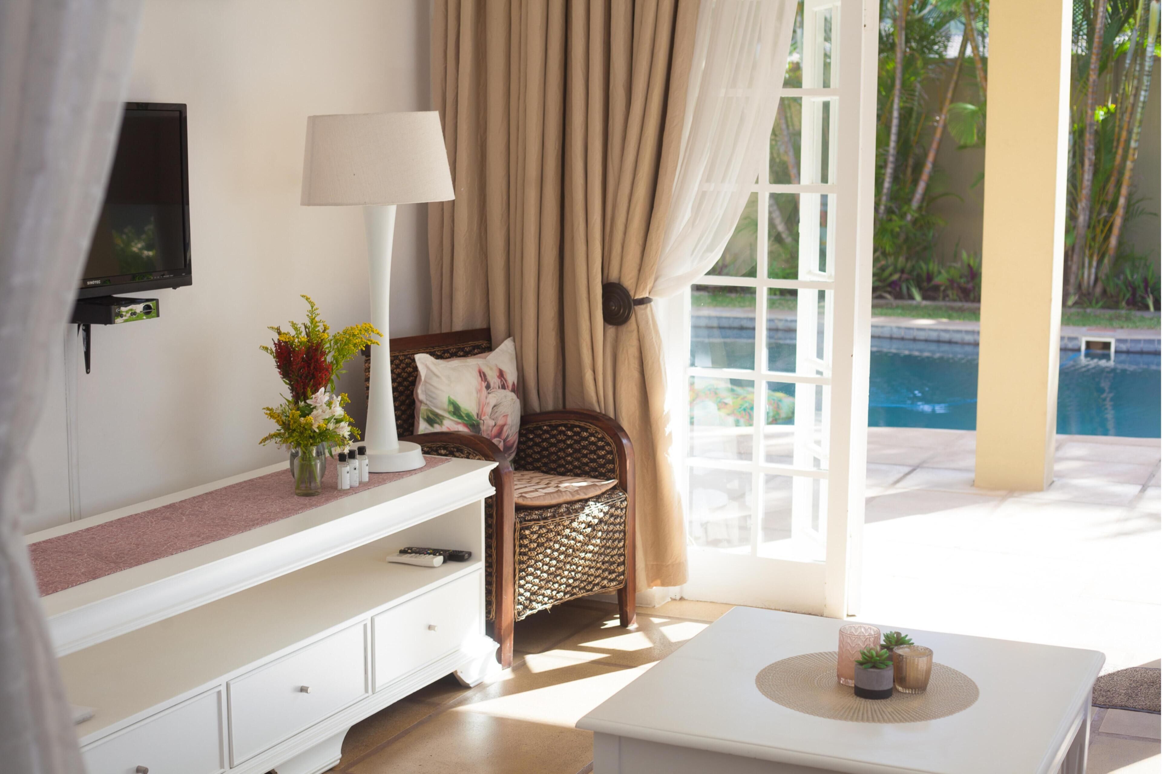A room with a white console table, a wicker chair with a cushion, a white armchair, a TV mounted on the wall, a lamp, and a view of a swimming pool.