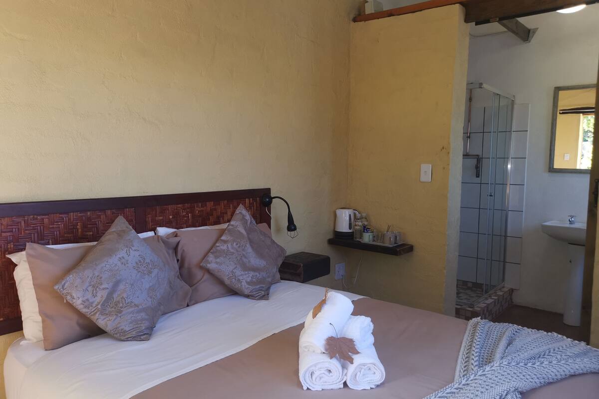 A neatly made bed with rolled towels and a blanket, a bedside table with a lamp, and a shelf with toiletries.