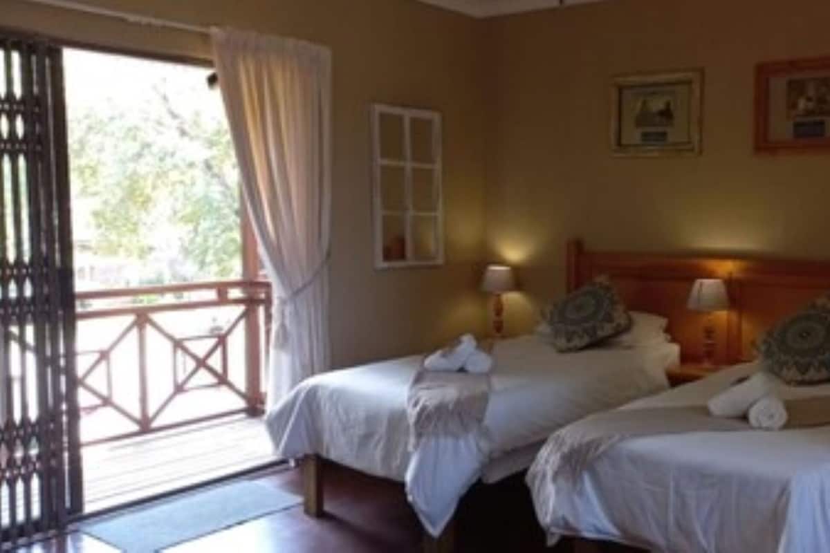 A room with two beds, a window with curtains, a wall-mounted mirror, and a small bathroom with a sink.
