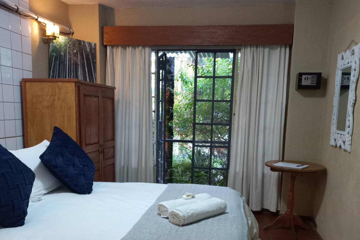 A neatly made bed with white linens and a grey herringbone throw blanket, a wooden wardrobe, a small round table with a mirror, and a window with curtains.
