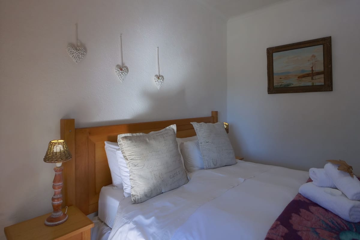 A neatly made bed with white linens and decorative pillows, a wooden headboard, a bedside table with a lamp, and a painting on the wall.