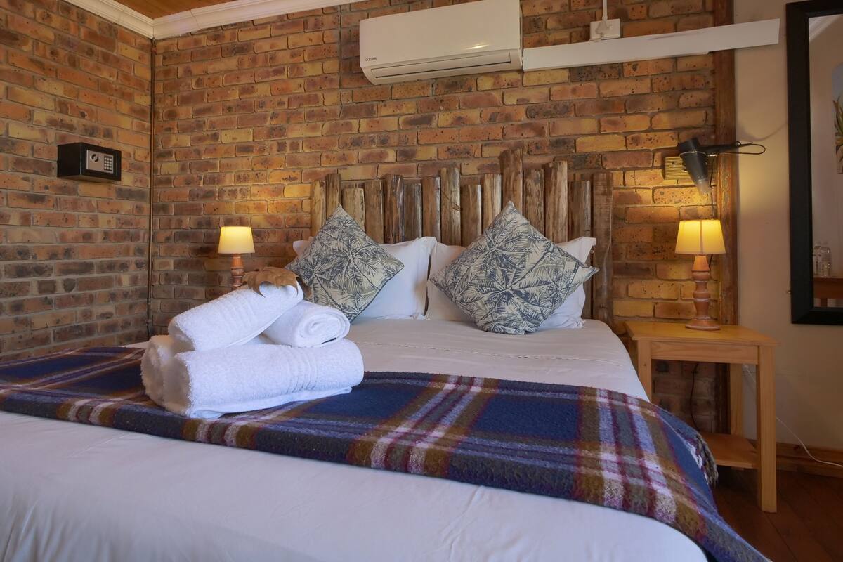 A hotel room with a brick wall, a bed with white linens, two pillows, a bedside table with a lamp, and an air conditioning unit.