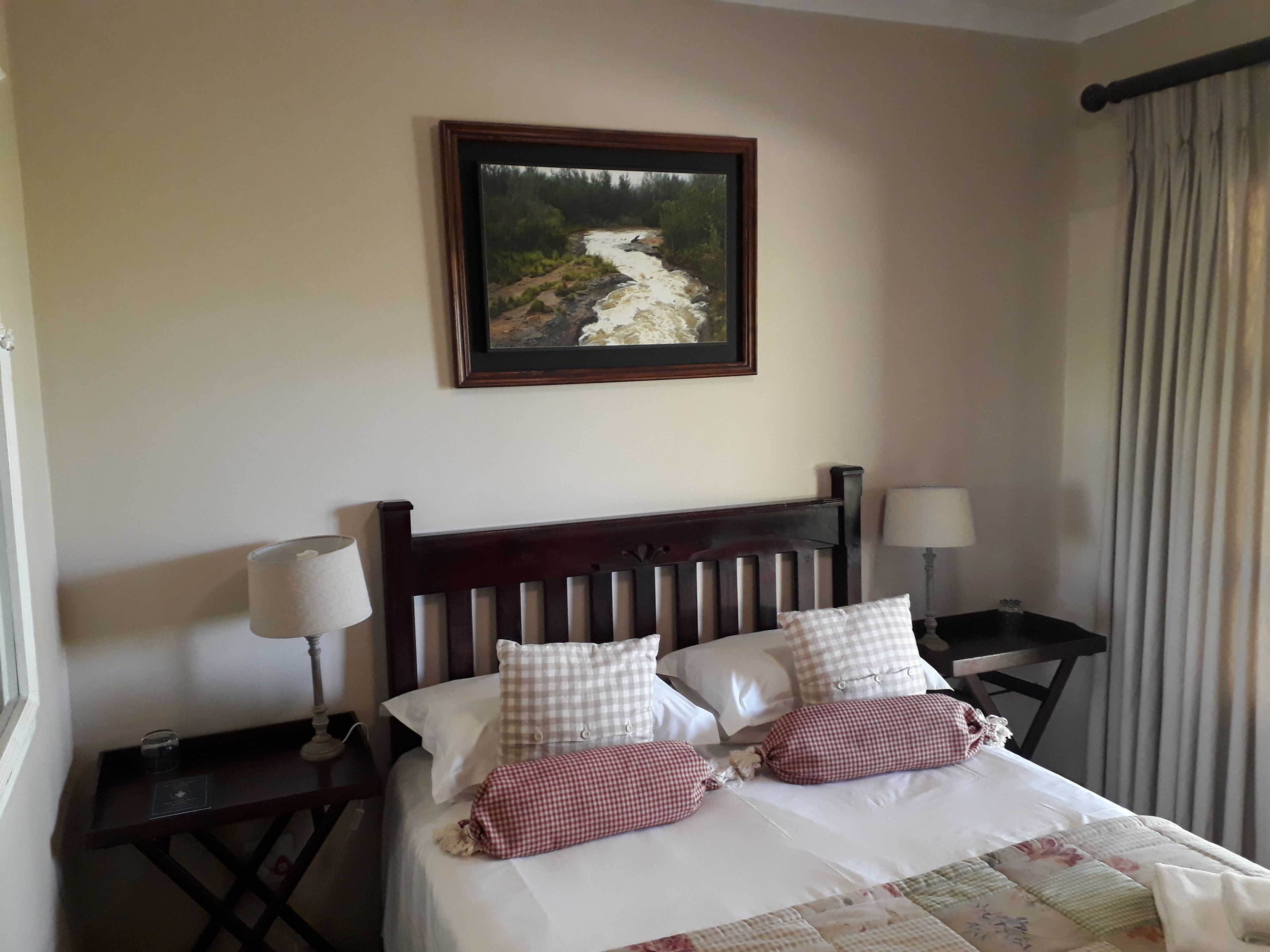 Una cama bien hecha con almohadones a cuadros, un cuadro enmarcado encima y una mesita de noche con una lámpara.
