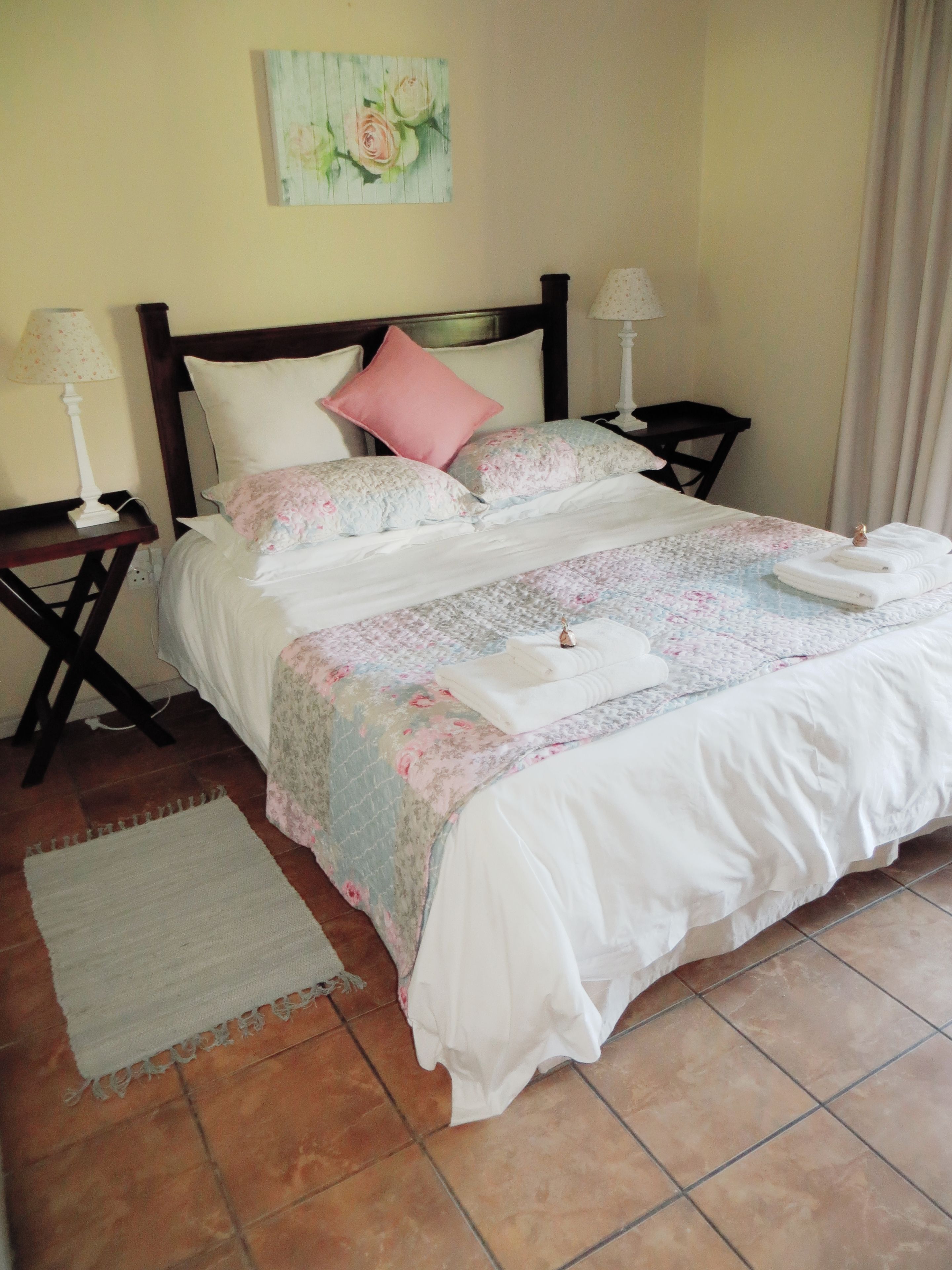 A neatly made bed with floral bedding, two bedside tables with lamps, a small rug, and a painting on the wall.