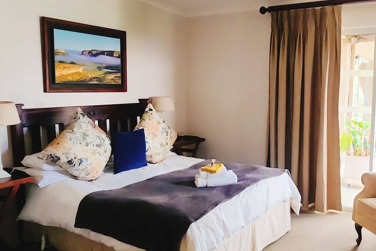 A hotel room with a bed, a chair, a lamp, a painting, and a view through the curtains.