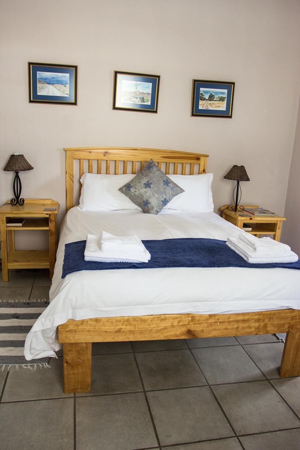 A neatly made bed with white linens, a blue throw blanket, and folded towels. There are two wooden nightstands with lamps, and three framed pictures on the wall.